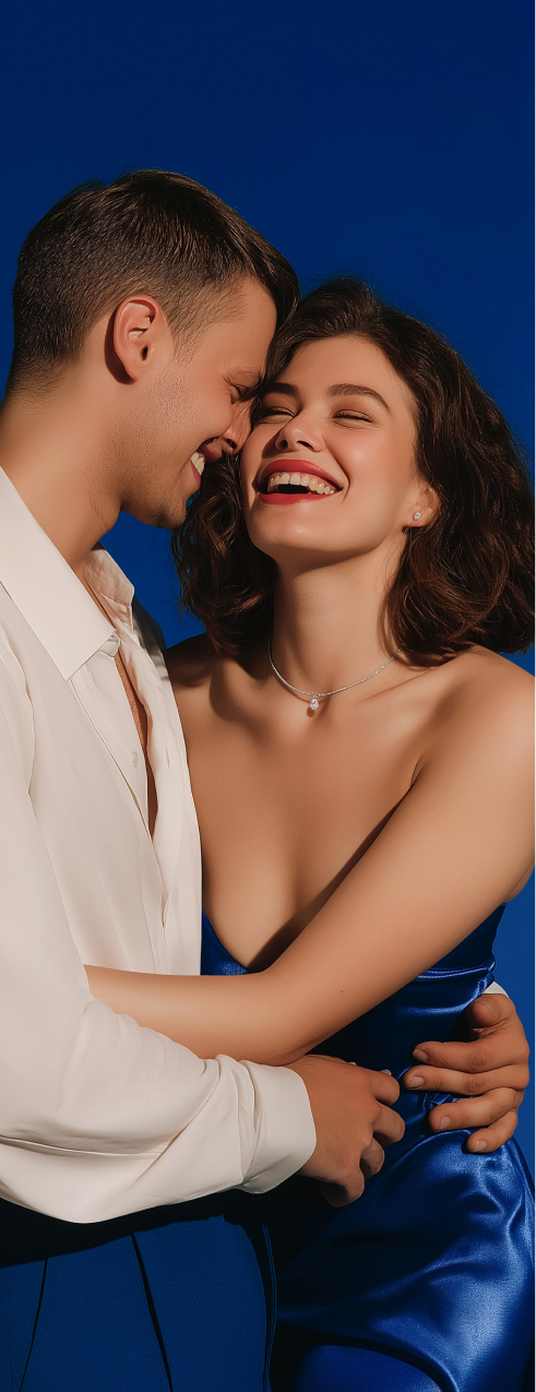 A young couple smiling on the blue backdrop, woman is wearing diamond jewelry.